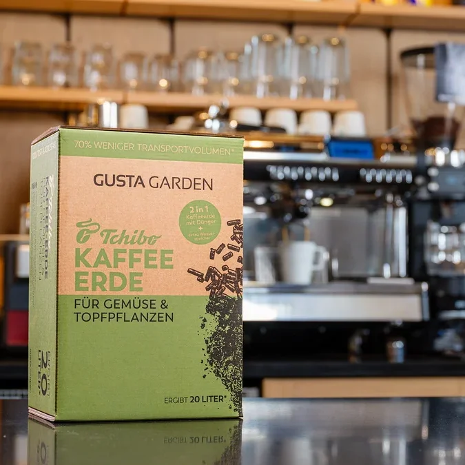 Eine Packung Tchibo Kaffee Erde steht vor einer Kaffeemaschine. Die Packung ist grün und braun und trägt die Aufschrift "Gusta Garden".
