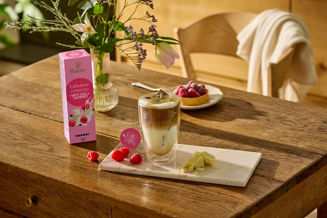 Auf einem Holztisch stehen ein Glas Kaffee, weiße Schokoladenstücke und Himbeeren auf einem Marmorbrett. Im Hintergrund ist eine Packung Cafissimo Flavoured Espresso - White Choc & Raspberry - 10 Kapseln zu sehen.