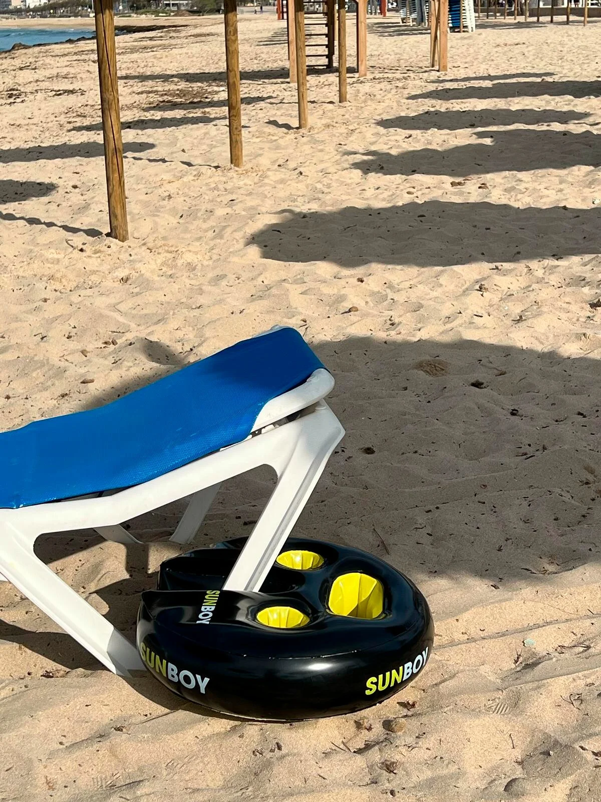 Auf einem Sandstrand steht ein Liegestuhl mit blauem Bezug auf einem schwarzen Sunboy Getränkehalter mit gelben Einsätzen.
