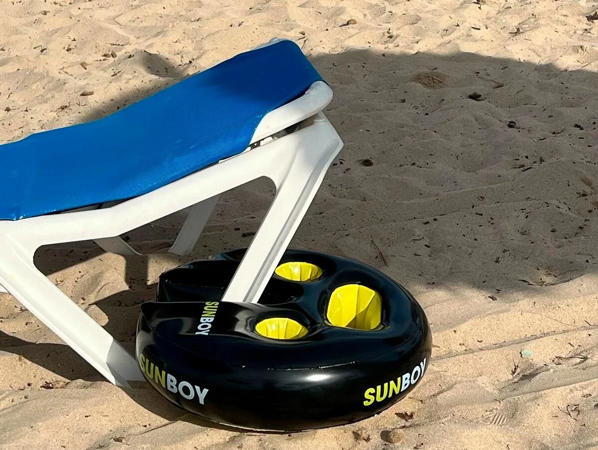 Auf einem Sandstrand steht ein Liegestuhl mit blauem Bezug auf einem schwarzen Sunboy Getränkehalter mit gelben Einsätzen.