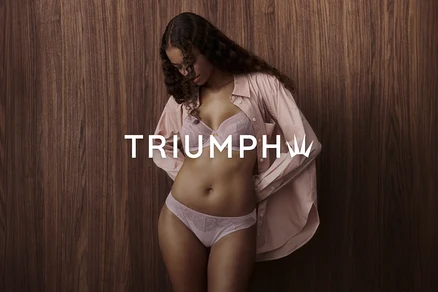 Eine Frau mit langen, lockigen Haaren trägt einen hellrosa BH und Slip sowie ein offenes, hellrosa Hemd vor einer Holzwand. Das Triumph-Logo ist sichtbar.