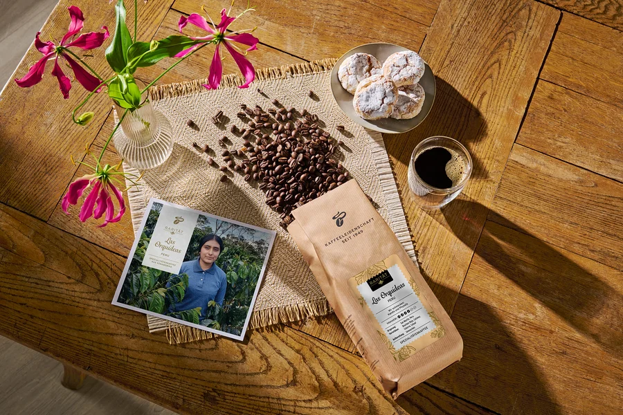 Auf einem Holztisch liegen eine geöffnete Packung Kaffeebohnen, daneben ein Glas Kaffee, ein Teller mit Gebäck, eine Postkarte und eine Vase mit Blumen.