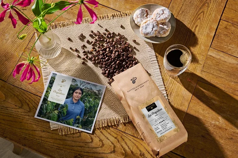 Auf einem Holztisch liegen eine geöffnete Packung Kaffeebohnen, daneben ein Glas Kaffee, ein Teller mit Gebäck, eine Postkarte und eine Vase mit Blumen.