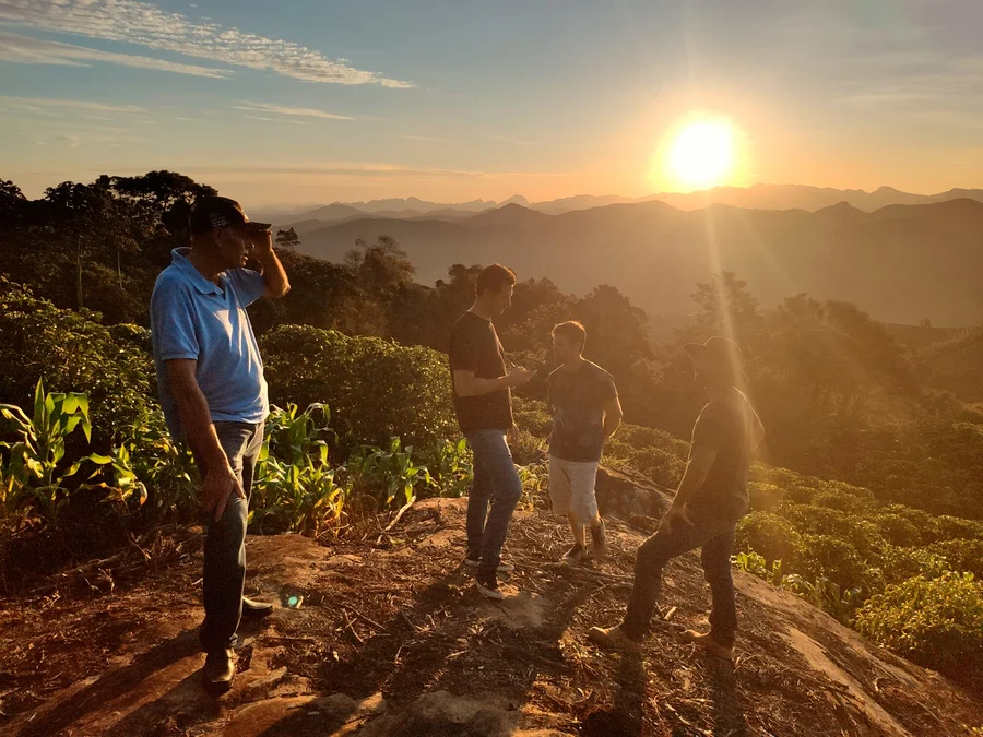 Vier Männer stehen auf einem Hügel bei Sonnenaufgang. Im Hintergrund sind Berge zu sehen.