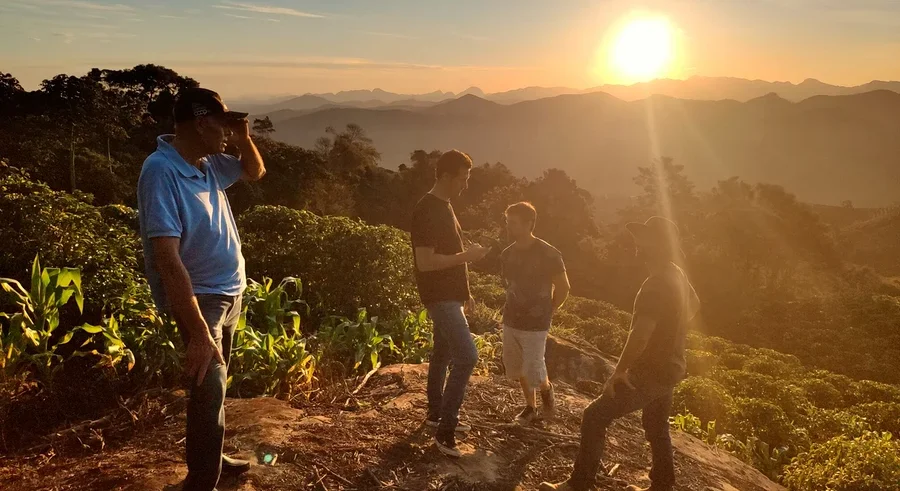 Vier Männer stehen auf einem Hügel bei Sonnenaufgang. Im Hintergrund sind Berge zu sehen.
