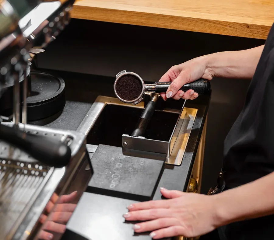 Eine Person hält einen Siebträger mit Kaffeepulver über einen Ausklopfkasten neben einer Kaffeemaschine.
