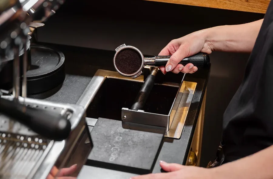 Eine Person hält einen Siebträger mit Kaffeepulver über einen Ausklopfkasten neben einer Kaffeemaschine.