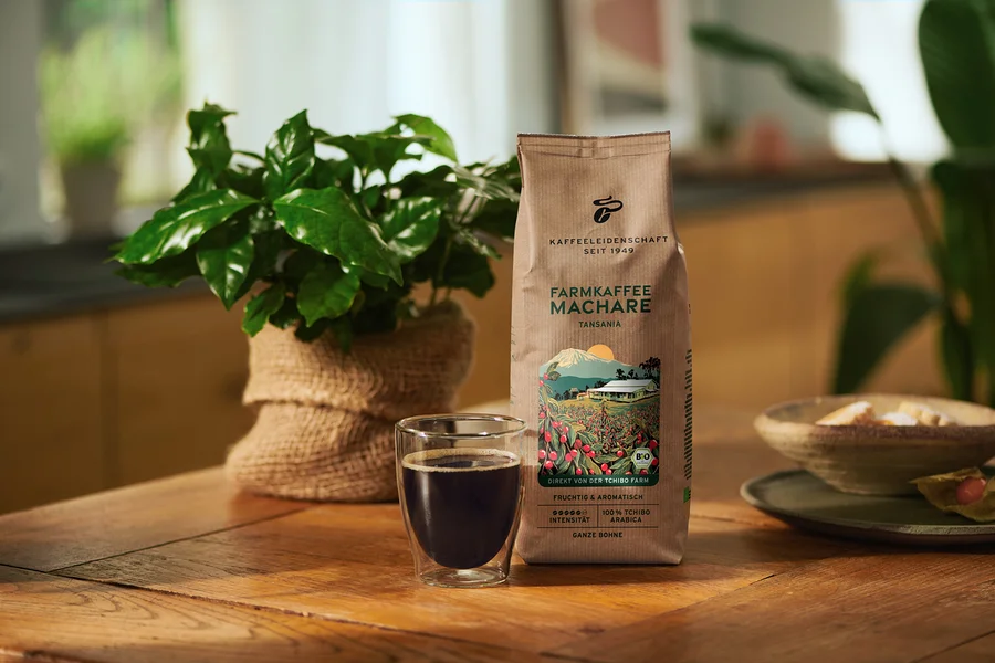 Braune Kaffeepackung "FARMKAFFEE MACHARE TANSANIA" neben einem Glas schwarzem Kaffee auf einem Holztisch, dahinter eine grüne Pflanze im Jutesack.
