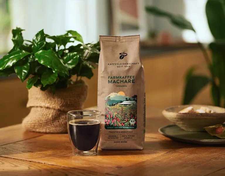 Braune Kaffeepackung "FARMKAFFEE MACHARE TANSANIA" neben einem Glas schwarzem Kaffee auf einem Holztisch, dahinter eine grüne Pflanze im Jutesack.