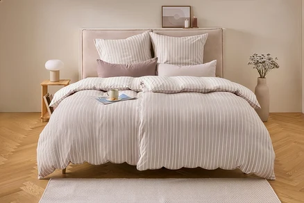 Ein Bett mit beiger und weiß gestreifter Flanell-Bettwäsche in Doppelgröße, darauf ein Magazin und eine Tasse. Links ein Nachttisch mit Lampe, rechts eine Vase mit Trockenblumen.