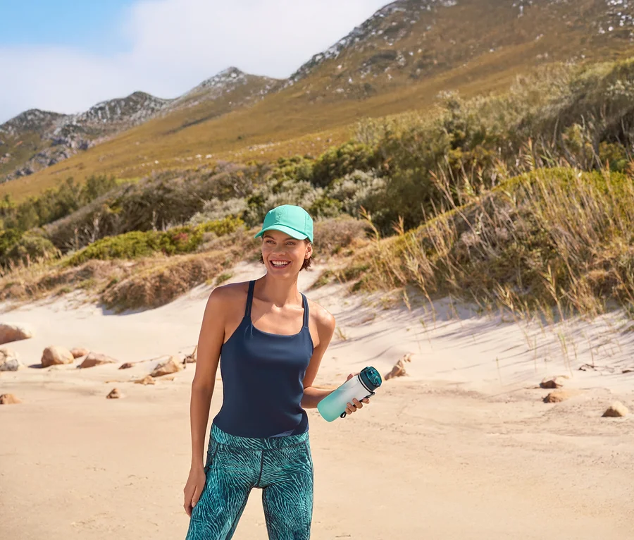 Lächelnde Frau in türkisener Sport-Kappe, dunkelblauem Yogatop mit integriertem BH und Sportradler »Active Power« mit Alloverprint, die eine Trinkflasche mit Kühlakku am Strand hält. Im Hintergrund sind Berge zu sehen.