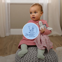 Ein Baby in einem rosa Kleid und hellgrünen Strumpfhosen sitzt auf einem grauen Pouf und hält ein graues Hasen-Stofftier mit einer rosa Decke. Im Hintergrund ist ein Zimmer mit Holzboden und einem Fenster zu sehen.