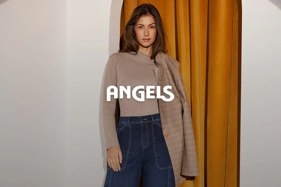 Eine Frau in einem beigefarbenen Langarmshirt und dunkelblauen Jeans, mit einem braun karierten Blazer über der Schulter, steht vor einer weißen Wand und einem goldenen Vorhang. Das Wort ANGELS ist überlagert.