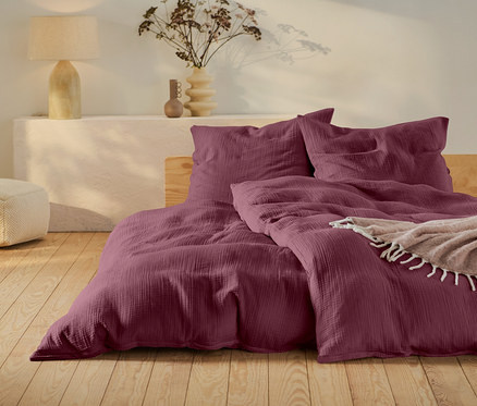 Weinrote Bettwäsche auf einem Bett mit Holzrahmen, ein beiger Pouf und eine Lampe auf einem weißen Tisch.
