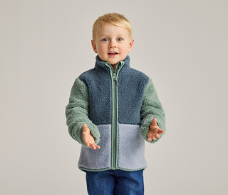 Ein blonder Junge mit blauen Augen trägt eine Fleecejacke in Blau, Grün und Hellblau und blaue Jeans.