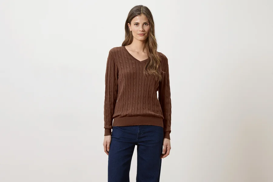 Frau mit langen braunen Haaren, die einen braunen V-Ausschnitt-Pullover und Jeans vor einem weißen Hintergrund trägt.