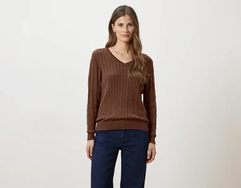 Frau mit langen braunen Haaren, die einen braunen V-Ausschnitt-Pullover und Jeans vor einem weißen Hintergrund trägt.