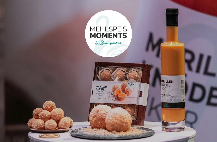 Auf einem Tisch stehen Marillenknödel Pralinen, Marillenknödel Likör und ein Logo von Mehlspeis Moments by Baumgartner.