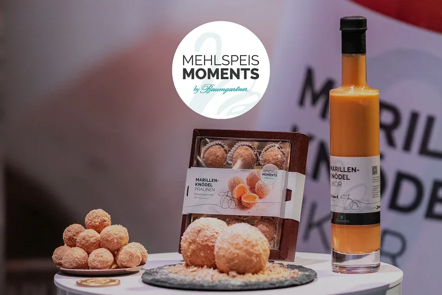Auf einem Tisch stehen Marillenknödel Pralinen, Marillenknödel Likör und ein Logo von Mehlspeis Moments by Baumgartner.