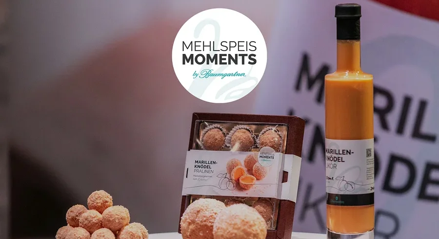 Auf einem Tisch stehen Marillenknödel Pralinen, Marillenknödel Likör und ein Logo von Mehlspeis Moments by Baumgartner.