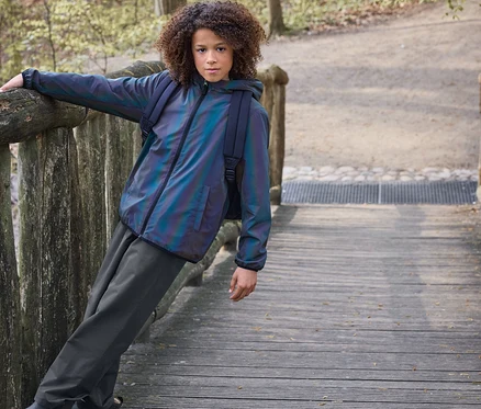 Kind mit Locken lehnt an Holzgeländer auf einer Brücke. Es trägt eine schillernde Jacke, dunkle Hose und braune gefütterte Kinder-Regen-Chelseaboots.