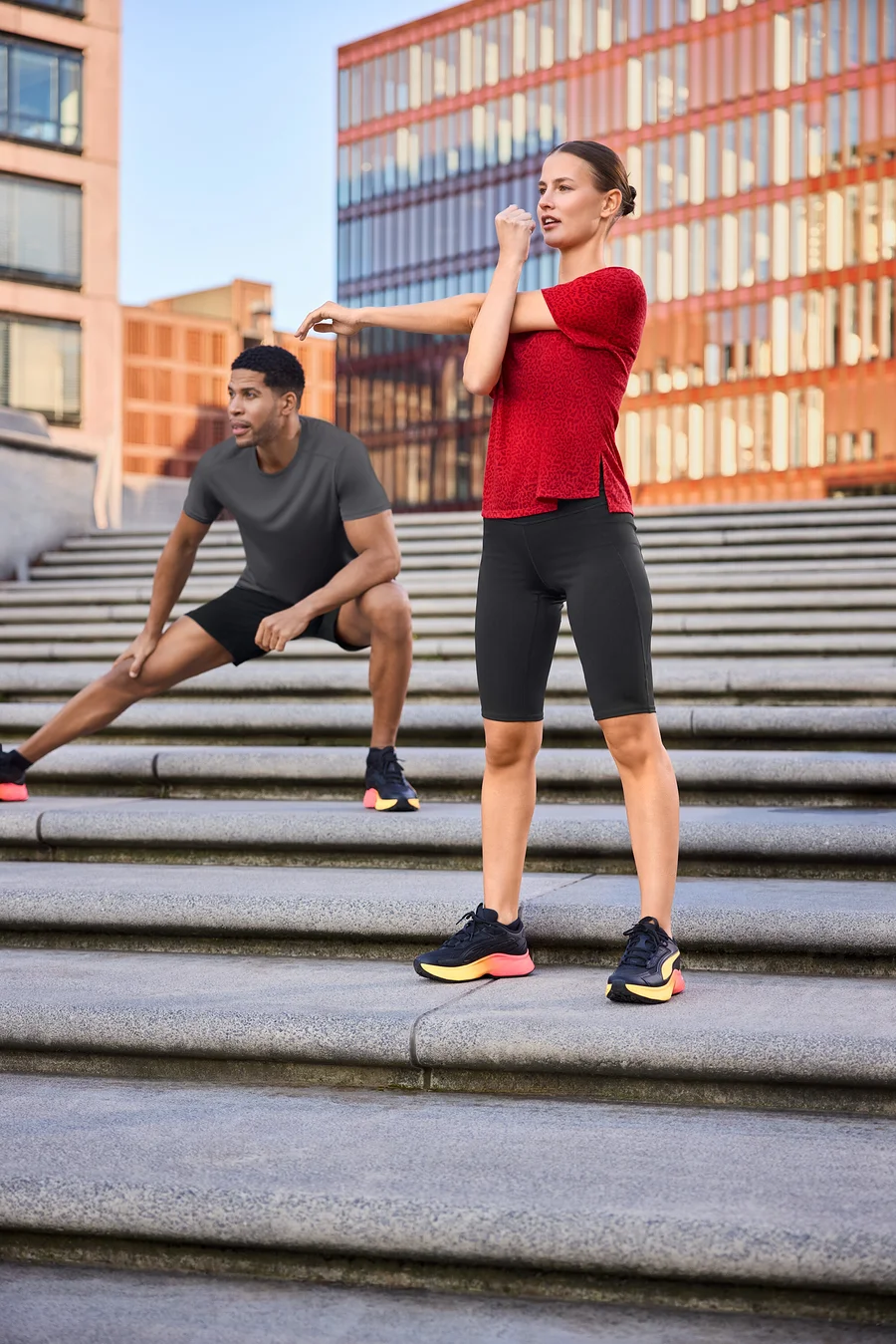 Frau in Sportradler und rotem Sportshirt dehnt sich auf Treppe, Mann in Sportshorts und grauem Funktionsshirt dehnt sich abseits.