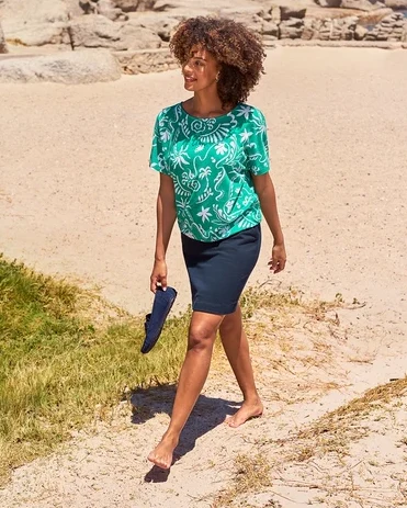 Barfuß geht eine Frau am Strand und hält Veloursleder-Loafer. Sie trägt Ohrstecker mit Glaskristallsteinen, einen Rock und ein grünes Oberteil.