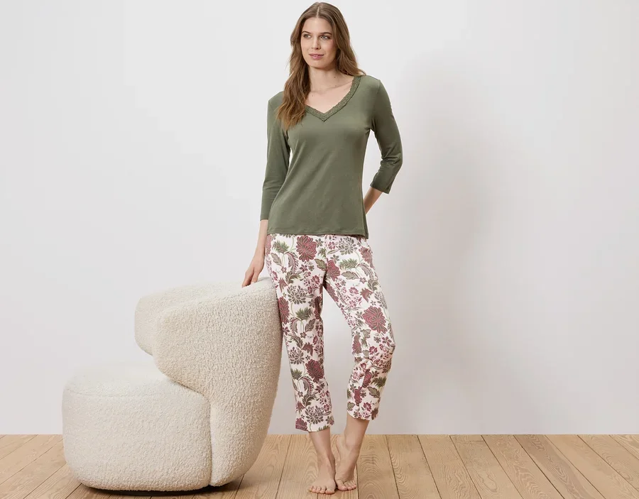Eine Frau steht neben einem Sessel und trägt einen olivgrünen Pullover und eine geblümte Hose.