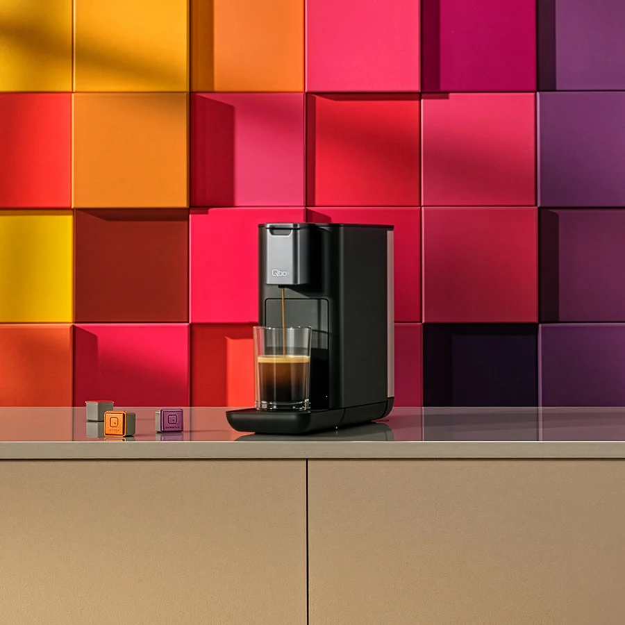 Eine schwarze Kaffeemaschine gießt Kaffee in ein Glas, mit zwei Kaffeekapseln daneben.