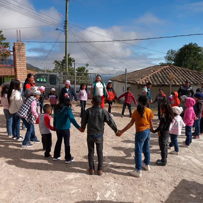 Eine Gruppe Kinder und Erwachsene hält sich im Kreis auf einem Feldweg im Freien an den Händen. Im Hintergrund sind ein rotes Tor, ein Lastwagen und ein Gebäude mit Ziegeldach zu sehen.