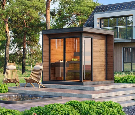 Eine hölzerne Außensaunakabine mit Glastüren steht auf einer erhöhten Betonplattform. Zwei hölzerne Liegestühle mit hellen Kissen und ein kleiner Whirlpool befinden sich auf dem angrenzenden Holzdeck. Im Hintergrund sind hohe Kiefern und ein modernes Haus mit Balkon zu sehen.