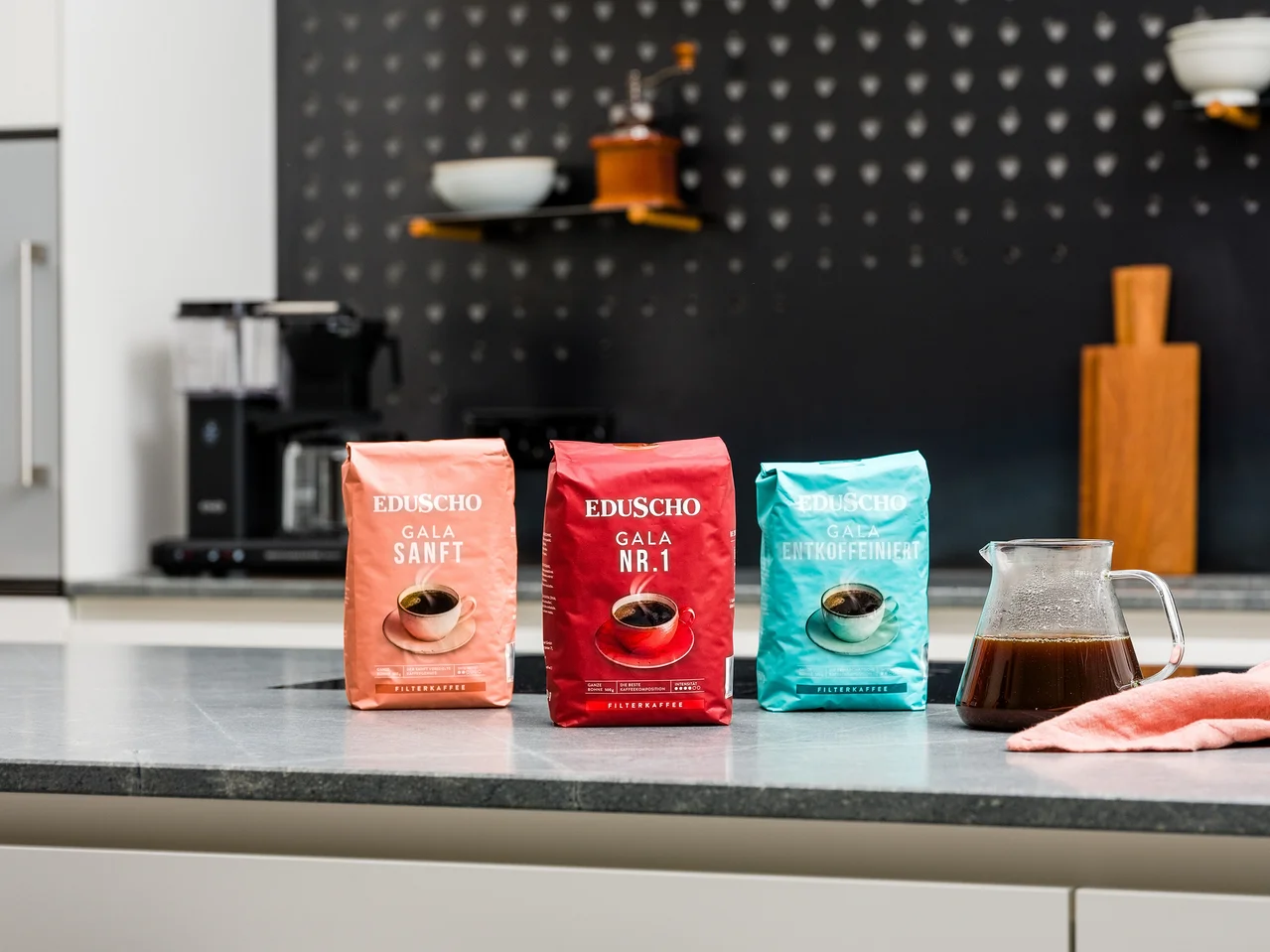 Drei Packungen gemahlener Kaffee von Eduscho Gala Sanft, Gala Nr. 1 und Entkoffeiniert stehen auf einer Küchentheke neben einer Glaskanne mit Kaffee.