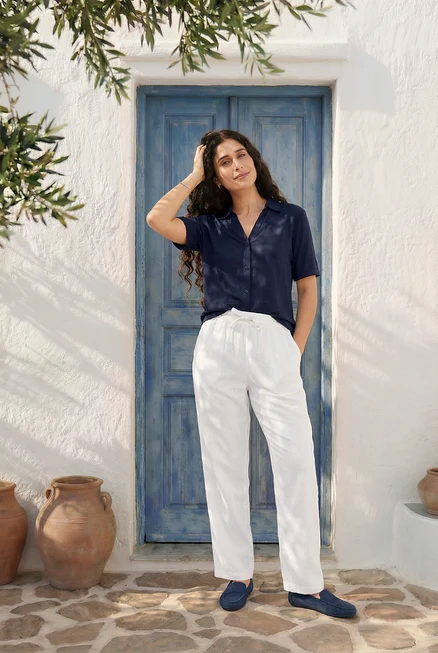 Frau posiert in blauem Blusenshirt mit Knopfleiste, weißer Leinenhose, Veloursleder-Loafern, Ohrsteckern und Tennisarmband vor einer blauen Tür.