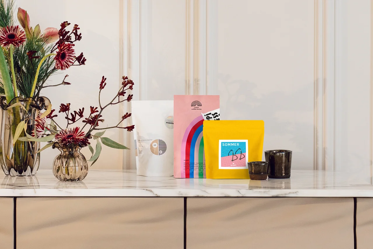 Drei bunte Produktbeutel in Weiß, Rosa mit Regenbogen und Gelb stehen auf einer Marmorarbeitsplatte neben zwei Vasen mit roten Blumen und zwei dunklen Tassen.