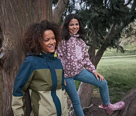Zwei lächelnde Mädchen in einem Park, eines trägt eine mehrfarbige Jacke, das andere sitzt auf einem Baumast in einer gemusterten Fleecejacke und Kinder-Slim-Jeans ‒ Fit »Lotta«.