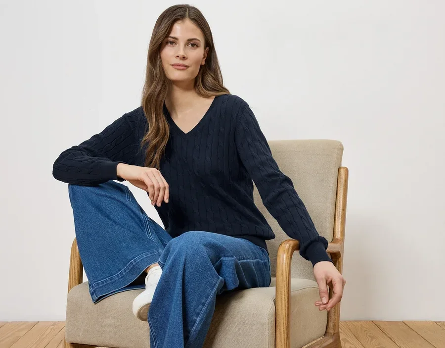 Frau sitzt in einem Sessel, trägt einen marineblauen V-Ausschnitt-Pullover und weite Jeans.