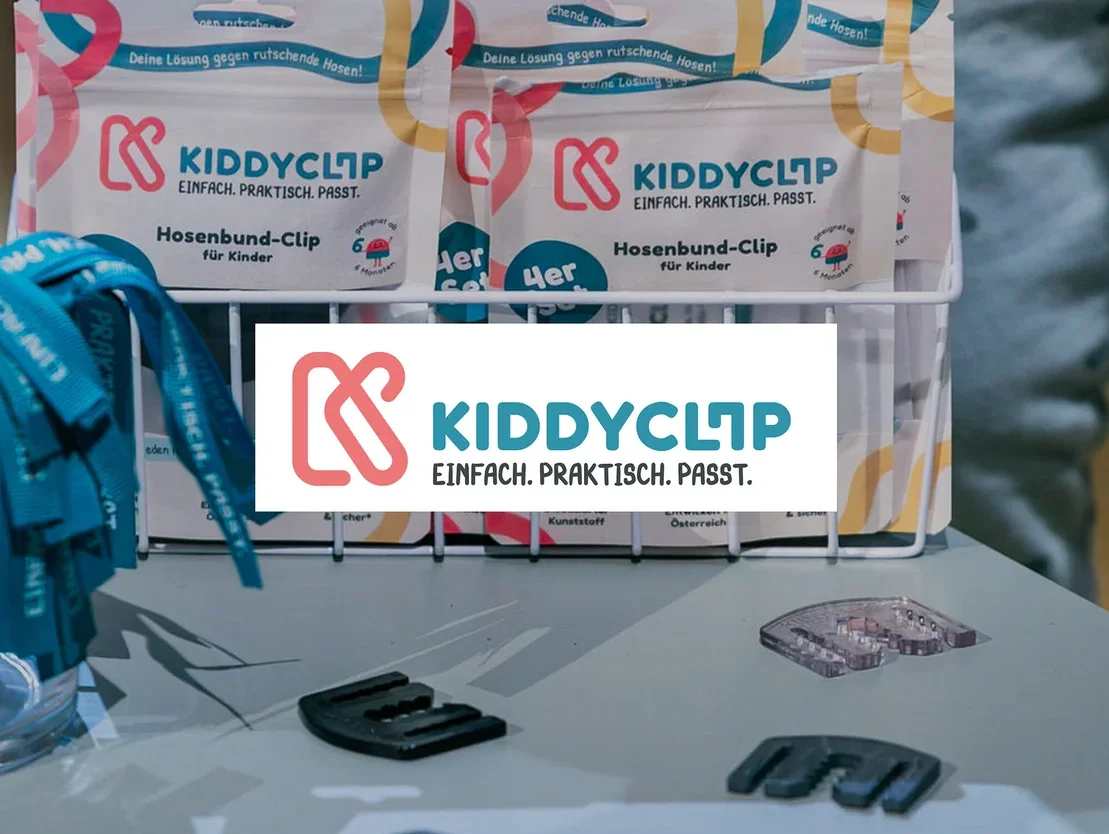 Auf einem Tisch stehen KiddyClips Hosenbund-Clips in Verpackungen und lose, daneben blaue Bänder in einem Glas.
