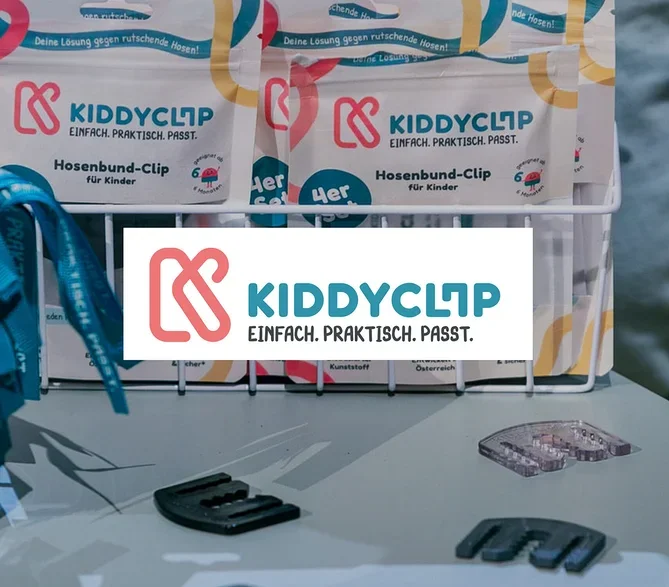 Auf einem Tisch stehen KiddyClips Hosenbund-Clips in Verpackungen und lose, daneben blaue Bänder in einem Glas.