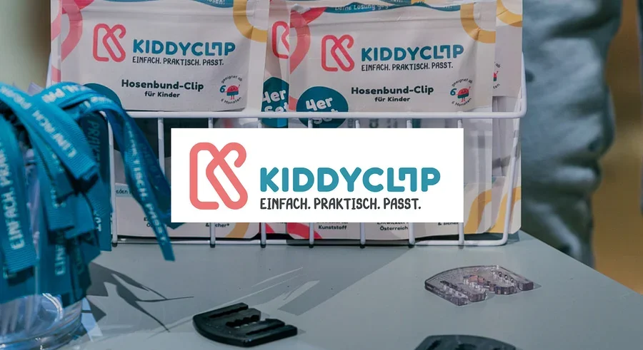 Auf einem Tisch stehen KiddyClips Hosenbund-Clips in Verpackungen und lose, daneben blaue Bänder in einem Glas.