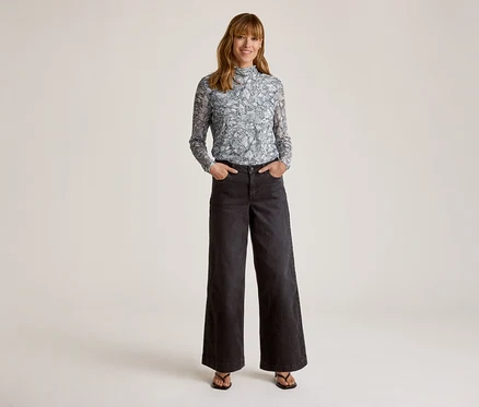 Lächelnde Frau in einem hellblauen, gemusterten Langarmshirt mit Stehkragen, dunklen weiten Hosen und dunklen Sandalen, die Hände in den Hosentaschen.