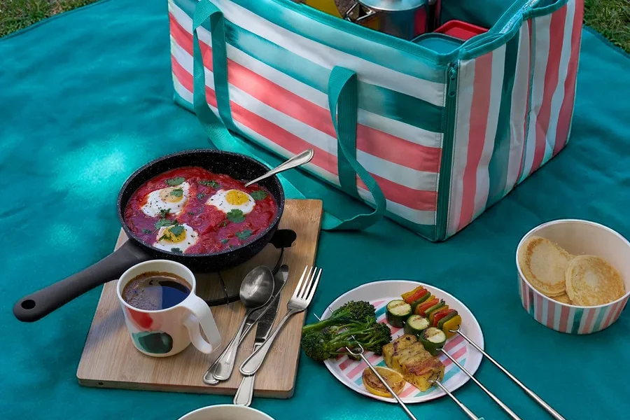 Auf einer Decke steht eine geöffnete Tasche neben einer Profi-Pfanne in Steinoptik (20 cm) mit Shakshuka, Geschirr, Grillspießen und Pfannkuchen.