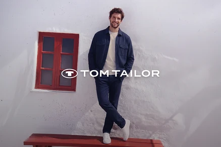Ein lächelnder Mann steht an einer weißen Wand gelehnt, trägt ein dunkelblaues Hemd, ein helles T-Shirt, dunkelblaue Hosen und weiße Sneaker. Seine Beine sind gekreuzt. Links von ihm ist ein rot gerahmtes Fenster, darunter eine rote Bank. Das Tom Tailor Logo ist sichtbar.