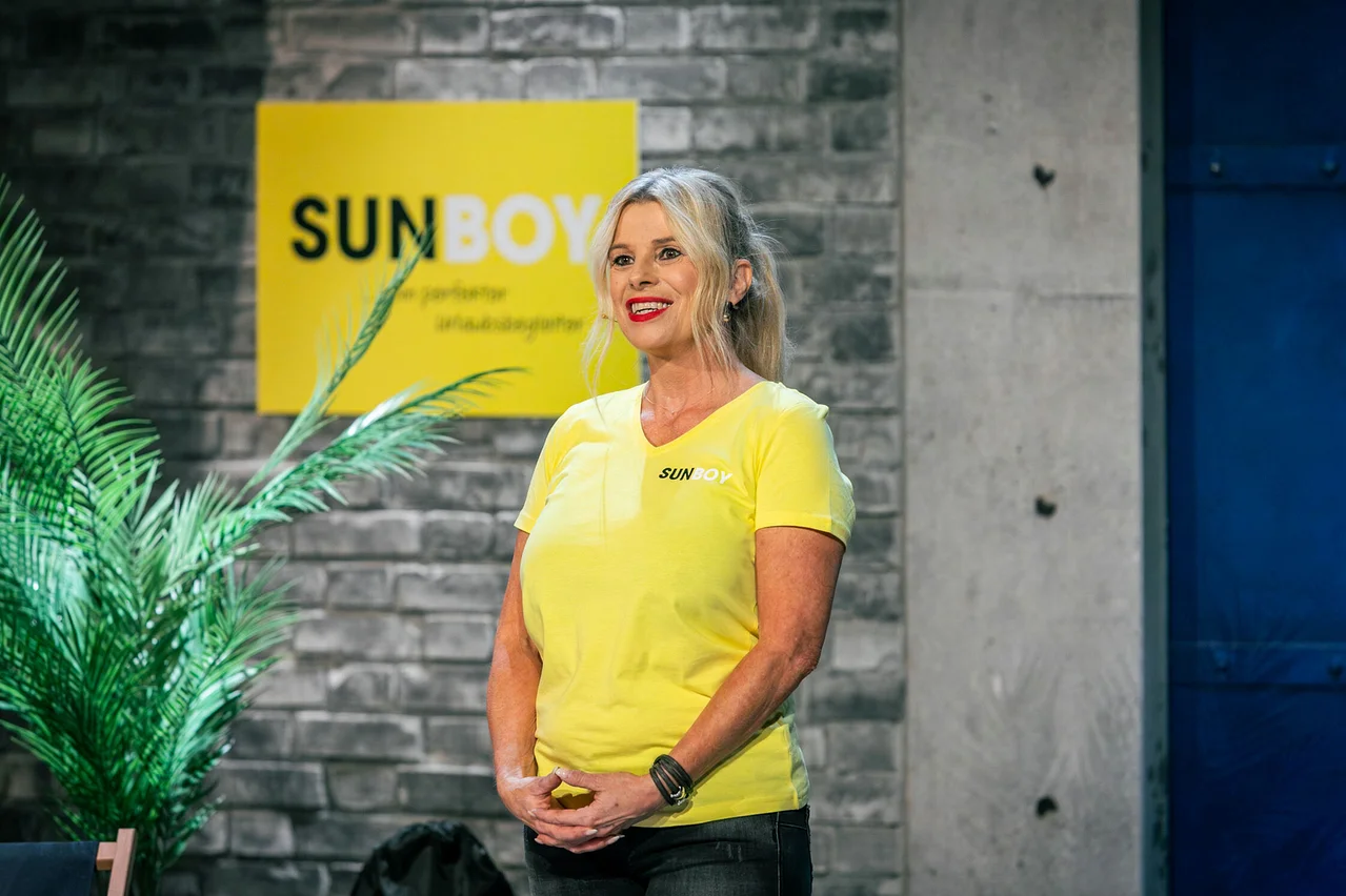 Frau mit blonden Haaren und gelbem T-Shirt steht vor einer Ziegelwand mit einem gelben SUNBOY-Schild.