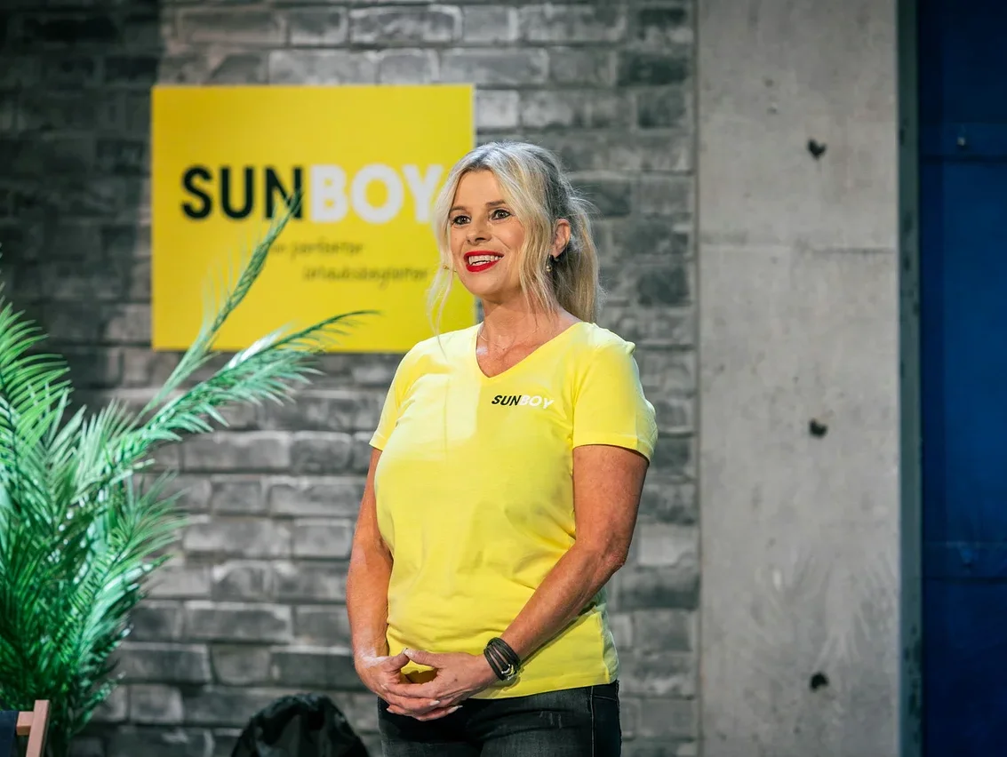 Frau mit blonden Haaren und gelbem T-Shirt steht vor einer Ziegelwand mit einem gelben SUNBOY-Schild.