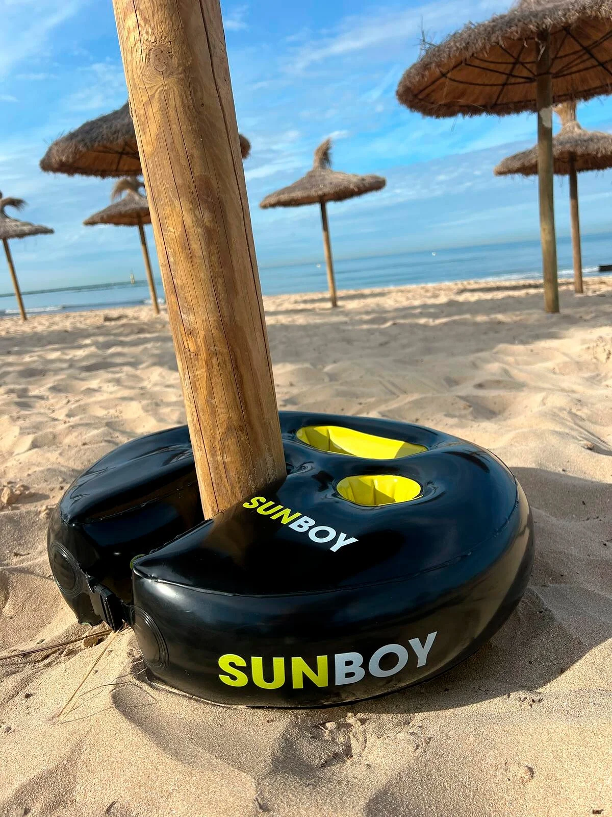 Ein Holzpfosten steckt in einem schwarzen SUNBOY-Ständer mit gelben Akzenten am Strand.