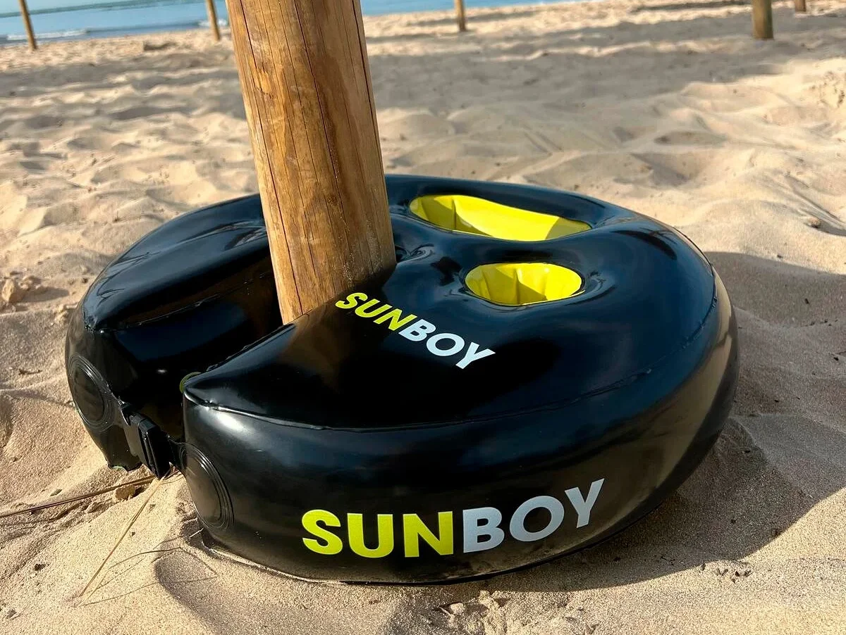 Ein Holzpfosten steckt in einem schwarzen SUNBOY-Ständer mit gelben Akzenten am Strand.