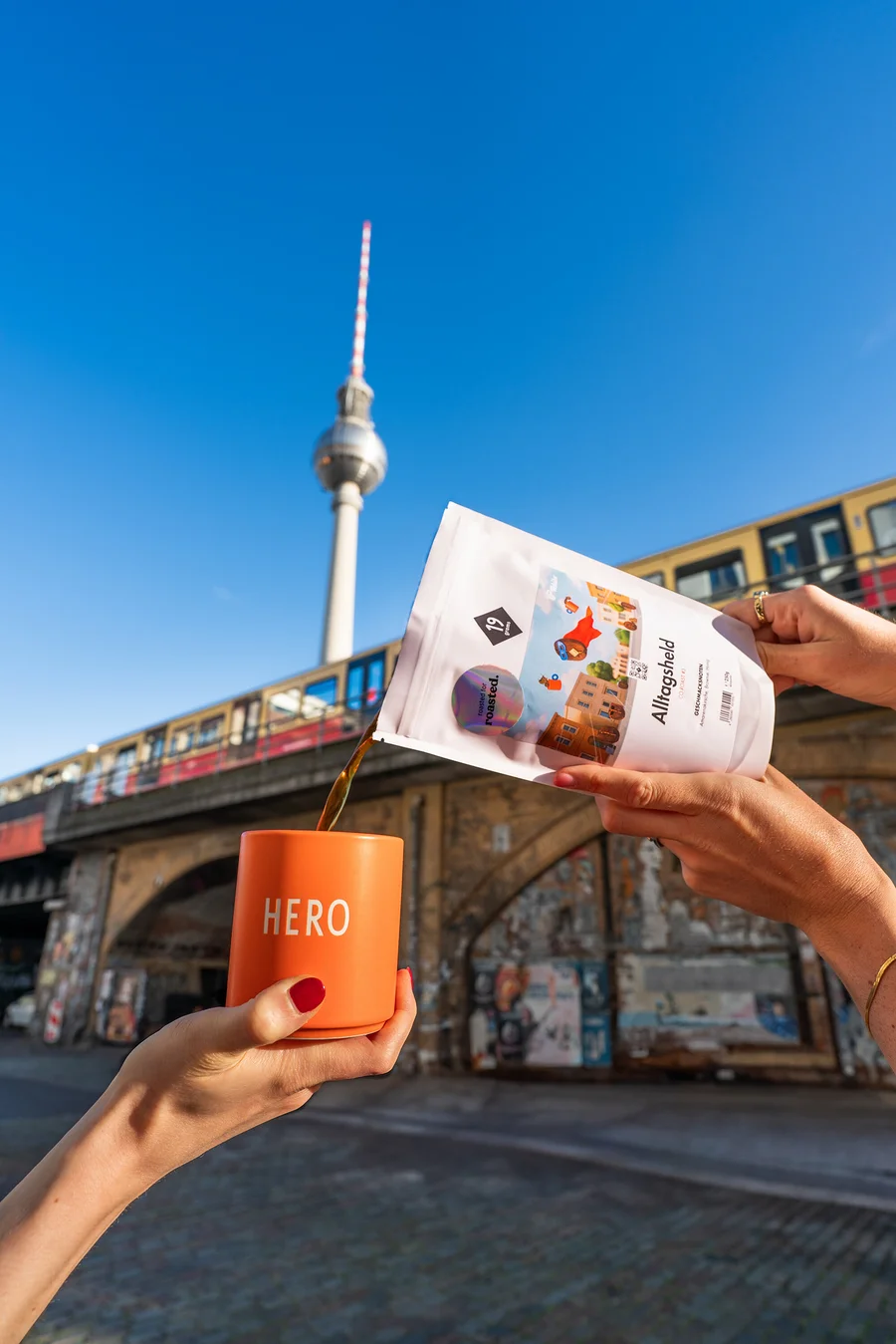 Eine Person gießt Kaffee aus einer weißen Packung in eine orangefarbene Tasse mit der Aufschrift HERO, im Hintergrund der Berliner Fernsehturm.