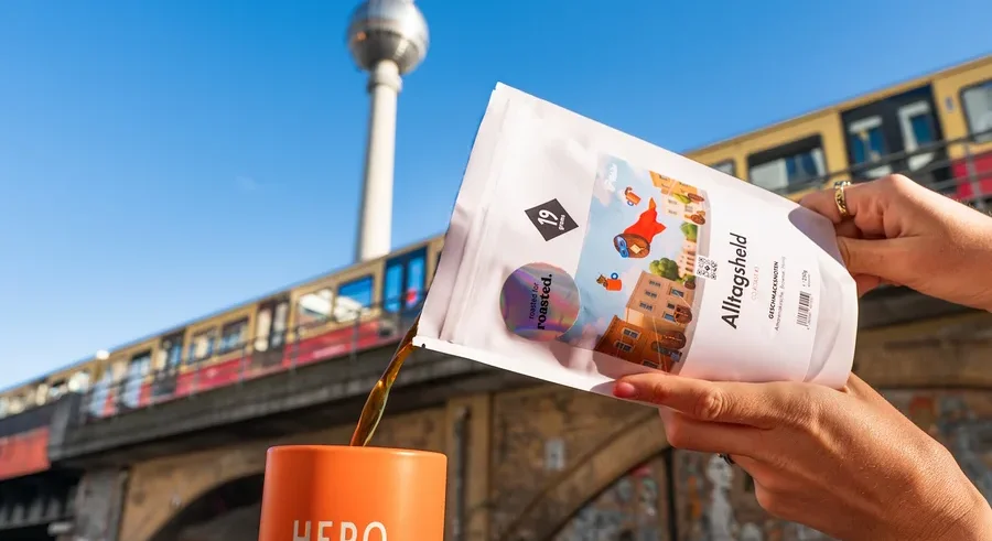 Eine Person gießt Kaffee aus einer weißen Packung in eine orangefarbene Tasse mit der Aufschrift HERO, im Hintergrund der Berliner Fernsehturm.