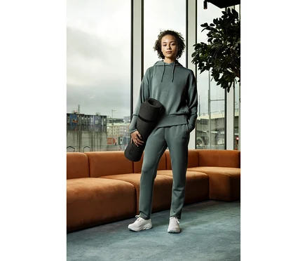Frau in einem salbeifarbenen FILA Damen-Hoodie aus Modalmix und passender Jogginghose, die eine schwarze Yogamatte hält, steht in einem modernen Raum mit großen Fenstern.