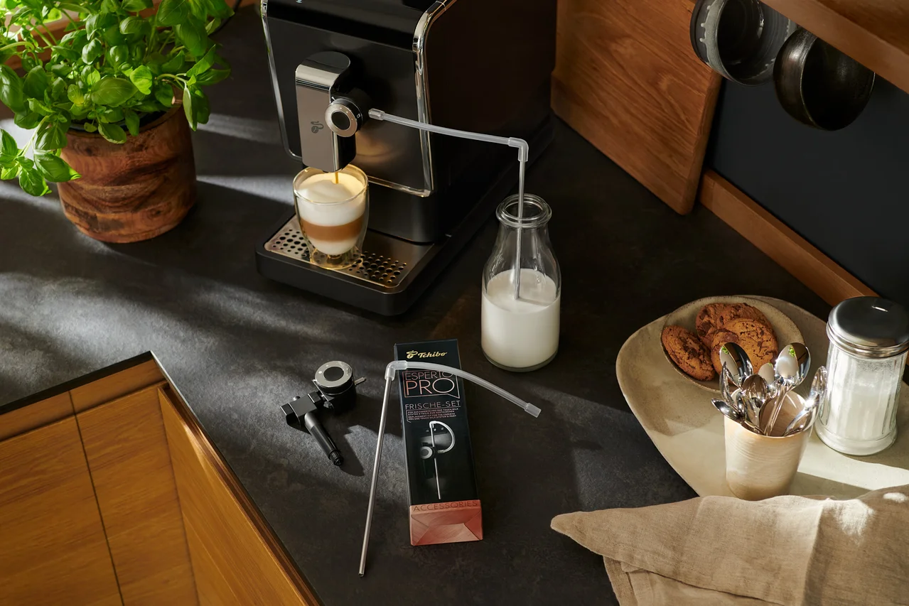 Ein »Esperto Pro« Tchibo Kaffeevollautomat bereitet Kaffee in einem Glas zu. Daneben stehen eine Milchflasche und Zubehör.